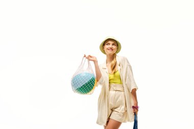 Şık yaz kıyafetleri içinde neşeli genç bir kadın neşeli bir plaj topu taşıyor ve gülümsüyor..