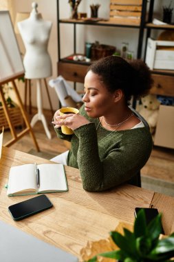 Bir zanaat atölyesinde, bir kadın düşünceli bir şekilde çayını yudumlarken etrafı malzeme ve fikirlerle çevrilidir..
