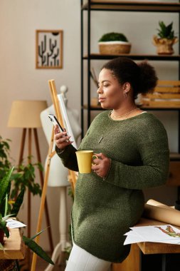 Güzel bir kadın, elinde sarı bir kupayla bir zanaat masasının yanında duruyor, telefonuyla meşgul..