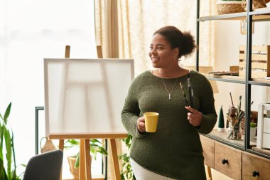 Güzel bir kadın, atölyesinin keyfini çıkarırken elinde bardak ve aletlerle gülümsüyor..
