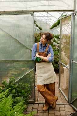 Genç bir kadın düşünceli bir şekilde büyüyen bitkilerle çevrili bir seraya yaslanır..