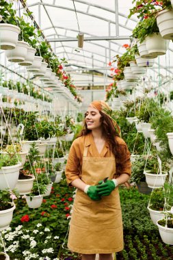 Başörtüsü ve önlüğü olan genç bir kadın yemyeşil bir serada bitkileri besliyor, mutluluk saçıyor..