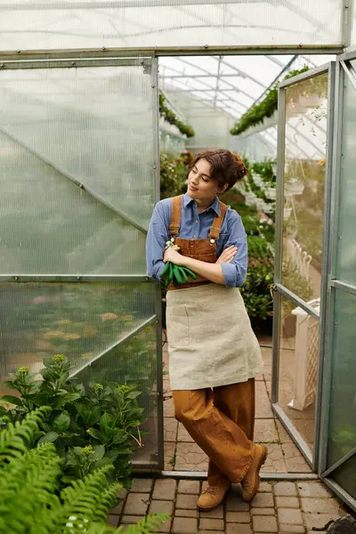 Genç bir kadın düşünceli bir şekilde büyüyen bitkilerle çevrili bir seraya yaslanır..