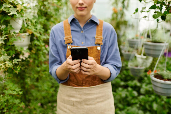 A young woman in an apron checks her phone amidst thriving plants in a greenhouse setting.