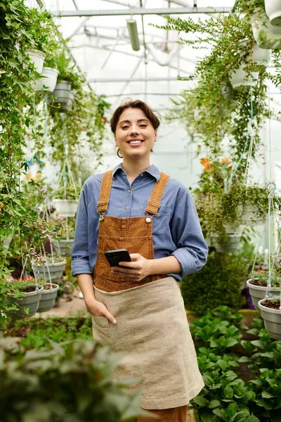 Neşeli bir bahçıvan parlak bir serada akıllı bir telefonu tutarken yemyeşil bitkileriyle ilgilenir..