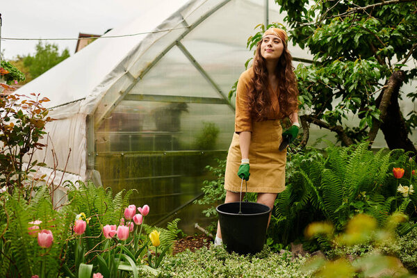 A young woman in a headscarf joyfully tends to plants in a vibrant greenhouse while wearing gloves.