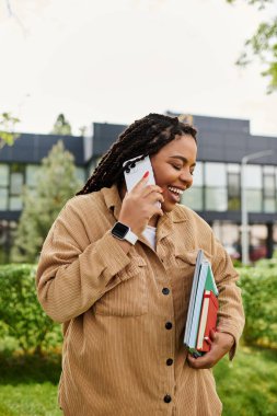 Neşeli bir üniversite öğretmeni, güneşli bir parkta kitap taşırken telefonla konuşur..