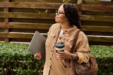 Sıradan giyinmiş bir kadın, elinde laptopuyla bir üniversite kampüsünün önünde yürüyor..