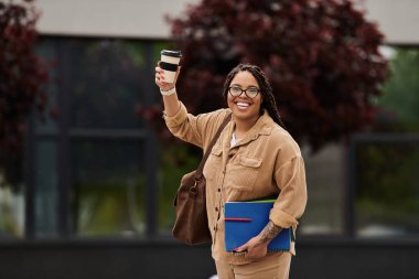 Neşeli üniversite öğretmeni elinde kahve tutarken ve akademik materyalleri dışarı taşırken el sallıyor.