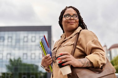 Bir üniversite öğretmeni elinde kitap ve kahve tutarken gülümsüyor, temiz havanın tadını çıkarıyor..