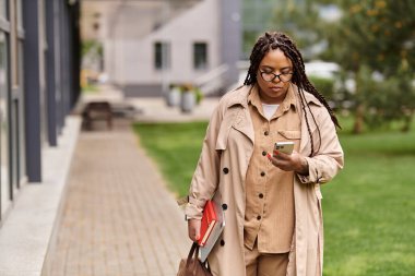 Bir üniversite öğretmeni şık bir trençkotla kampüs yollarında dolaşıp telefonunu kontrol ediyor..