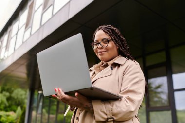 Trençkotlu bir kadın üniversite binasının önünde laptopunu kullanırken gülümsüyor..