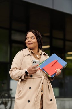 Bir üniversite öğretmeni elinde renkli defterler ve akıllı bir telefonla dışarıda durup gülümsüyor..