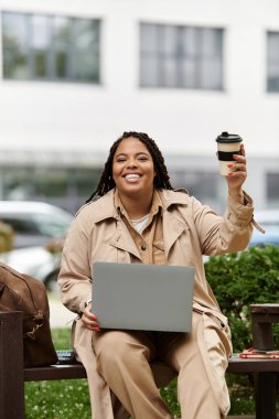 Üniversite eğitimcisi bankta oturur, kahveyle gülümser ve dizüstü bilgisayarda yazar..