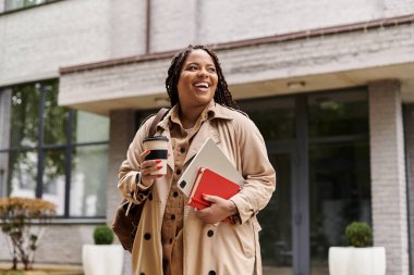 Gülümseyen üniversite öğretmeni kampüste yürürken elinde kahve fincanı ve kitaplar tutuyor..