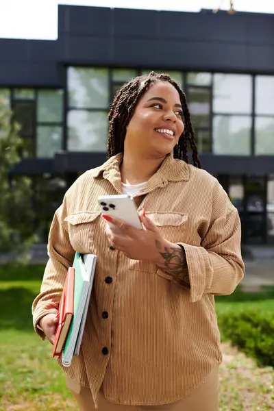 Kendine güvenen bir üniversite öğretmeni kampüste gülümsüyor, telefonuyla meşgul oluyor ve elinde kitap tutuyor..