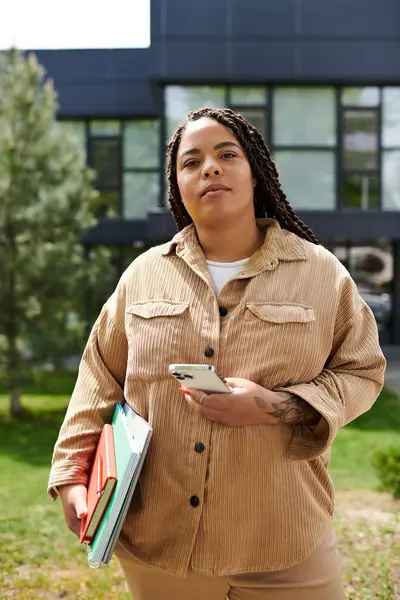 Kendini işine adamış bir öğretmen elinde defterler ve akıllı bir telefonla güvenle dışarıda duruyor..