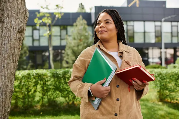 Bir üniversite öğretmeni sınıfların yanında yeşil bir alanda renkli defterlerle gülümser..