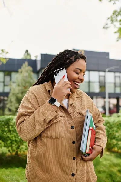 Neşeli bir üniversite öğretmeni, güneşli bir parkta kitap taşırken telefonla konuşur..