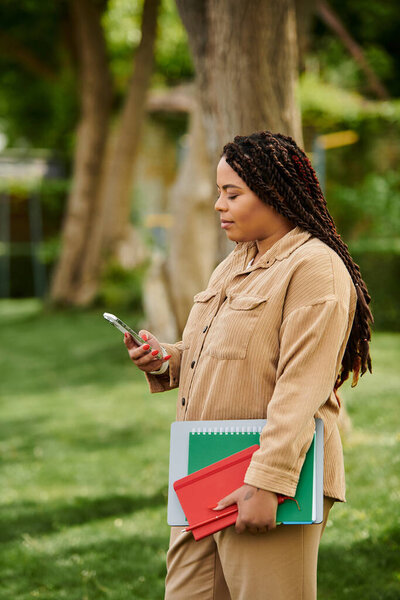 Учительница университета стоит на улице, проверяя смартфон, балансируя книги на руках..