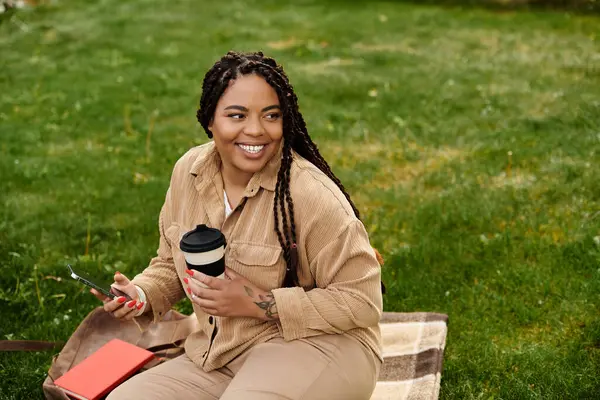Üniversite öğretmeni çimenli bir alanda rahatlar, kahvenin tadını çıkarır ve gülümserken telefonunu kullanır..
