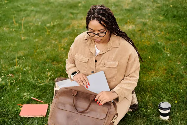 Bir kadın çimenlerde oturur ve üniversite sınıfına hazırlanırken dizüstü bilgisayarını kullanır ve notlar alır..