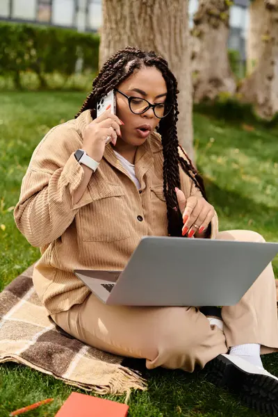 Yeşillik bir park alanında dizüstü bilgisayarla çalışan bir kadın telefonda konuşuyor..