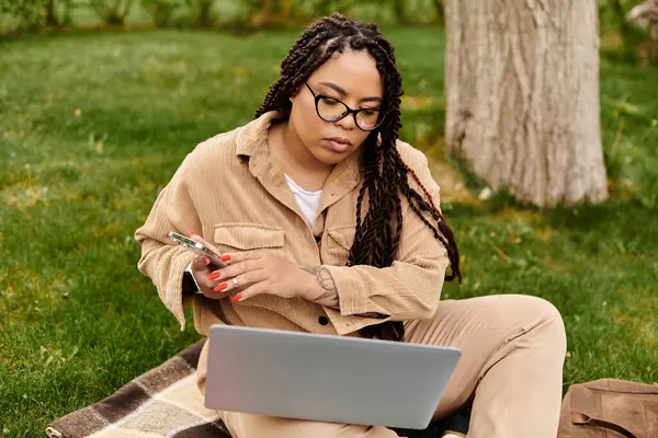 Bir kadın çimenlere rahatça oturur, dizüstü bilgisayar ve akıllı telefon kullanır, işine odaklanır..