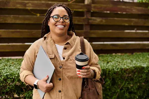 Bir kadın eğitmen, kampüste dizüstü bilgisayar ve kahve fincanı tutarken güneşli bir günün tadını çıkarır..