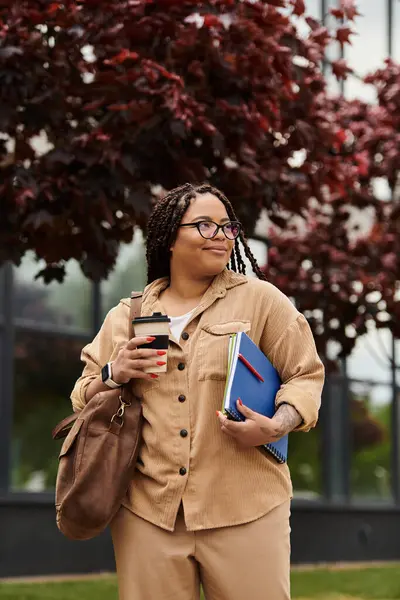 Bir üniversite öğretmeni elinde kahve fincanı ve kitap tutarken güzel bir yeşilliğin yanında durur..
