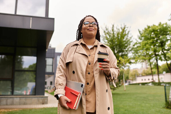 A university teacher walks confidently across the lush campus holding coffee and a tablet, smiling.