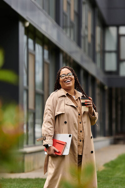 A joyful woman in a tan coat holds coffee and a notebook, surrounded by university buildings.