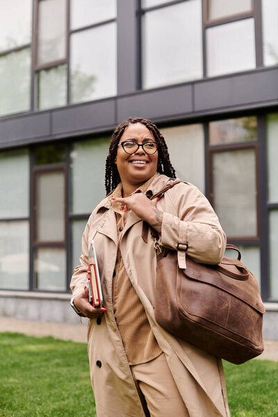 Woman in a light coat smiling while holding books and a bag on university grounds.