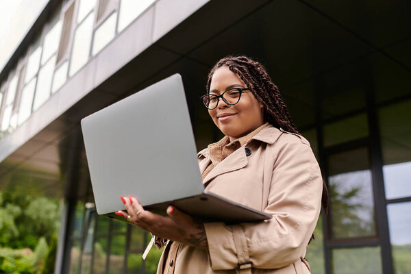 Женщина в плаще улыбается, когда пользуется ноутбуком возле здания университета..