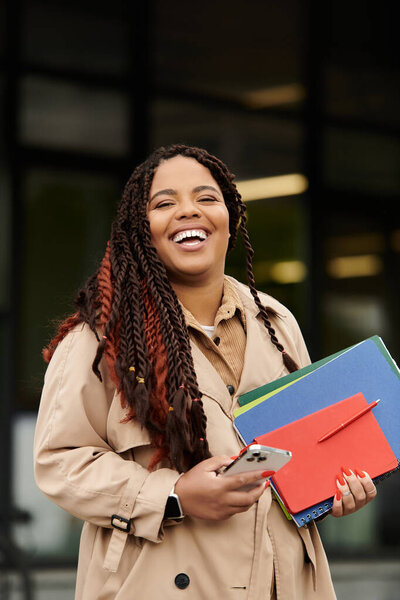 A woman educator stands with happiness, holding colorful binders and a phone near her campus.