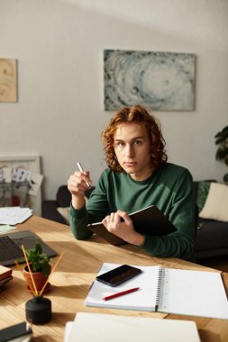 Kıvırcık saçlı genç bir adam ahşap bir masada oturur ve fikirlerini not defterine yazar..