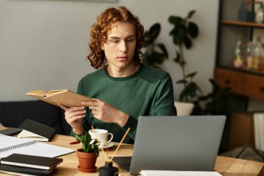 Genç bir adam rahat bir şekilde ev ortamında oturup dizüstü bilgisayarını kullanarak kitap okuyor..