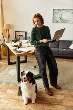 Genç bir adam, köpeği yakınlarda dinlenirken rahat bir evde masa başında çalışıyor..