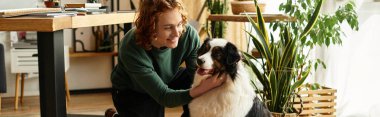 Genç bir adam diz çöker, gülümser ve sıcak, davetkar bir yaşam alanında köpeğini okşar..