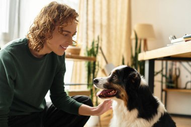 Genç bir adam parlak ve rahat bir oturma odasında köpeğiyle oynarken sıcak bir şekilde gülümsüyor..