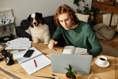 Genç bir adam ahşap bir masada çalışırken, bir köpek de rahat bir evde merakla izliyor..