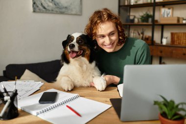Genç bir adam ahşap bir masada oturuyor, internette köpeğiyle konuşurken gülümsüyor..
