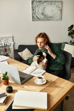 Genç bir adam ahşap bir masada oturuyor, köpeği yanına sokulurken telefonla konuşuyor..