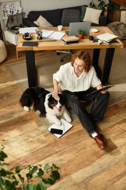 Genç bir adam rahatça yerde oturuyor, köpeğiyle meşgul oluyor ve notlar yazıyor..