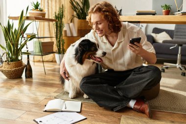 Genç bir adam köpeğiyle yerde oturuyor, telefonda planlarını düzenlerken gülümsüyor..