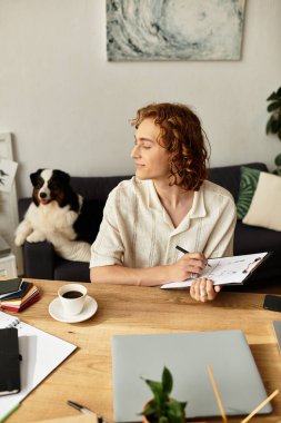 Genç bir adam ahşap masasında rahatça oturur, köpeği yakınlarda dinlenirken çizim yapar..