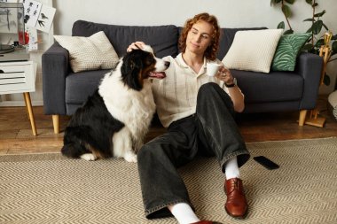 Genç adam kahvesini yudumlarken ve evde köpeğini okşarken rahatça yerde oturuyor..