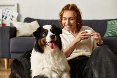 Genç bir adam yerde rahatça oturup kahvesini yudumlarken neşeli köpeği yakınlarda dinleniyor..