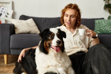 Genç bir adam elinde kupayla neşeyle yerde oturuyor ve köpeğiyle vakit geçiriyor..