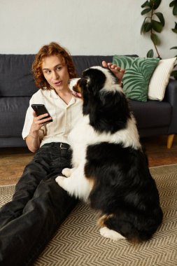 Genç bir adam rahat oturma odasında yerde oturuyor, köpeği ve akıllı telefonuyla meşgul..